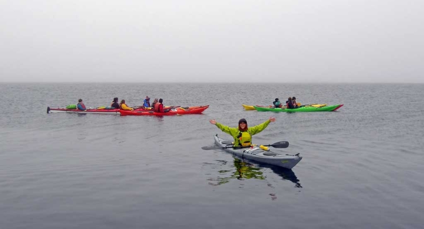 Colorful kayaks are paddled by students on calm water under gray skies. 